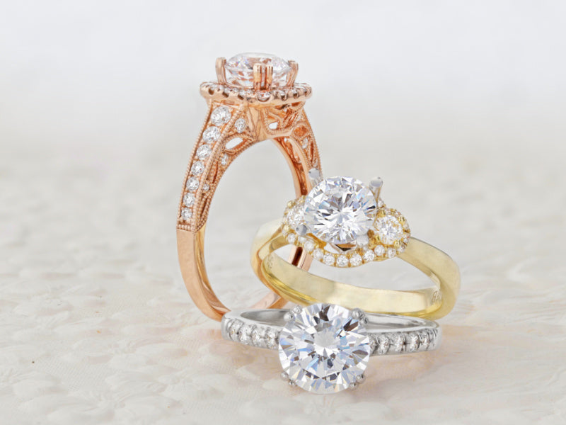 A rose gold ring with a halo, diamonds on the shank, and ornate details; a yellow gold three stone halo ring; a white gold ring with diamonds on the side and double prongs.