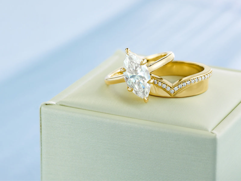 A gold engagement ring with a diamond marquise-cut center stone and a matching gold band with small pavé stones, displayed on a cream jewelry box against a soft light blue background.