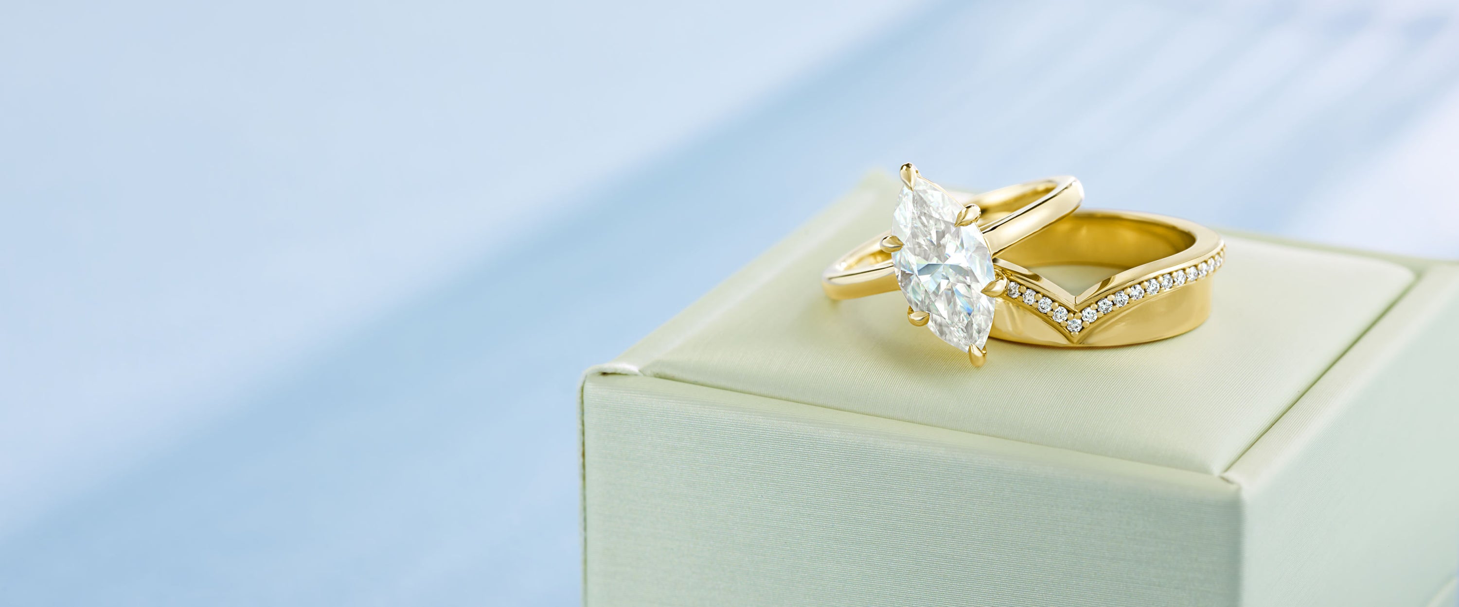 A gold engagement ring with a diamond marquise-cut center stone and a matching gold band with small pavé stones, displayed on a cream jewelry box against a soft light blue background.